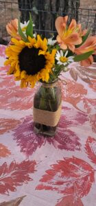 A small flower arrangement in a glass jar with a jute twine wrap, featuring sunflowers, orange lilies, and white daisies, sits on a pink and red leaf-patterned tablecloth outdoors, reminiscent of an art gallery display.