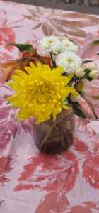 A small bouquet in a glass vase adorns a table with a floral-patterned cloth, much like an exhibit in a gallery. The bouquet features a bright yellow chrysanthemum, white daisy-like flowers, an orange lily, and some greenery with white berries.