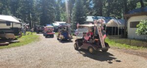 A festive parade of decorated golf carts drives down a gravel path in a wooded campground, resembling a moving gallery. The carts are adorned with American flags and patriotic decorations. Campers and trailers are parked nearby, and a few people stand watching the parade.