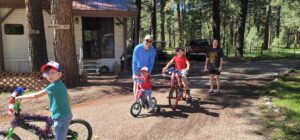A group of people outdoors near a forest and a house resembles a lively gallery. Two adults stand while three children ride decorated bicycles. One adult bends down to assist a child. The area is shaded with trees, and a pathway leads away from the house.