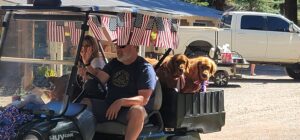A man drives a golf cart decorated with American flags, accompanied by a woman waving and two brown dogs. The cart is outdoors on a sunny day, with a truck and a building visible in the background, resembling an open-air gallery of small-town Americana.