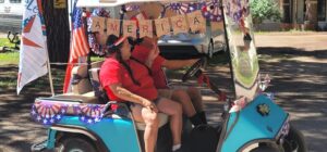 Two people in red shirts ride in a blue golf cart decorated for Independence Day, adorned with "America" and patriotic themes in red, white, and blue. They appear to be participating in a festive event or parade, reminiscent of a fine art tableau, surrounded by trees and parked vehicles.