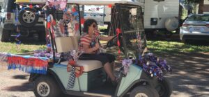 A woman drives a golf cart decorated with red, white, and blue streamers, ribbons, and stars that suggest a patriotic celebration. She wears a striped shirt and sunglasses. The background looks like a lively gallery with an RV and a few parked cars adding to the scene.