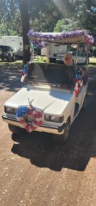 A white golf cart adorned with red, white, and blue decorative garlands and ribbons. An elderly person with short white hair and sunglasses is driving the golf cart, creating a scene as striking as an art gallery exhibition. Trees, parked vehicles, and a campground area are visible in the background.