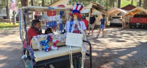 A golf cart adorned with red, white, and blue decorations and tinsel at a campground. Two people sit in the cart, while a mannequin dressed in patriotic attire holds a sign reading, "Tall Wild City Camp Site Mascot, Feel Me!" It feels like an art gallery showcasing festive creativity outdoors.