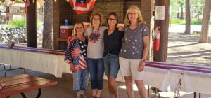 Four women standing under a pavilion decorated with American flags are smiling, dressed in patriotic-themed clothing, and have their arms around each other. Like a lively gallery of Americana, a red and white striped tablecloth lines the table behind them, along with a cooler and a fire extinguisher.