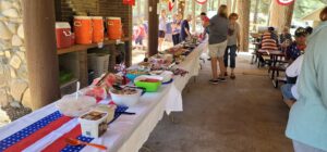 A buffet table is set up outdoors at a pavilion, featuring a variety of foods including watermelon, salads, and desserts. Amidst the American flag-themed decorations and tablecloths, the setup almost resembles an exhibition space where people gather around to serve themselves.