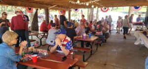 People gathered under a wooden pavilion for a picnic event, which felt like an outdoor gallery. Some are seated at red picnic tables, chatting and eating, while others stand nearby. The area is decorated with patriotic banners and bunting. The atmosphere is lively, and there are food tables in the background.