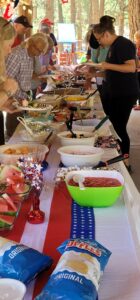 People are gathered around a long, buffet-style table filled with various dishes and snacks. The table, reminiscent of an art exhibition, is decorated with a red, white, and blue theme, including a flag-themed tablecloth. There are bags of chips, bowls of fruit, and other food items on display.