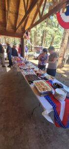 A long table is set up under a wooden pavilion, resembling an outdoor gallery of culinary delights, with a variety of dishes for the picnic. Two women are selecting food. The pavilion is adorned with patriotic bunting in red, white, and blue. People are gathered around, and a wooded area is visible in the background.