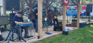 A group of musicians performs outdoors on a wooden stage, which resembles a vibrant gallery. One man is playing a pedal steel guitar while the others play electric guitars and drums. A painted backdrop with "Saddleback" decorates the stage. Trees and a grassy area are visible in the background.