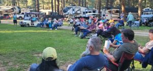 A large group of people, seated in folding chairs and relaxing on the grass, are gathered outdoors at a picturesque campsite with several RVs and trailers in the background. Tall pine trees surround the area like a natural gallery, and some people are watching an unseen speaker or event to the left.