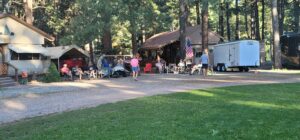 A group of people enjoying a campsite with RVs, a trailer, and a small cabin surrounded by trees. Some are sitting in chairs, others standing or walking, creating a lively outdoor gallery. An American flag is displayed near one of the structures.
