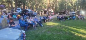 A group of people sitting on lawn chairs in a grassy area surrounded by tall trees, giving the scene a fine art quality. There are several parked vehicles and cabins in the background. The sunlight filters through the trees, casting shadows on the relaxed crowd as if it were an open-air exhibition.