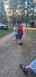 Two people are dancing on a gravel path outdoors, surrounded by trees and a few parked vehicles, including a truck and an RV. The person on the left, in a light blue shirt and denim shorts, twirls with flair, while the one on the right sports a red shirt, black shorts, and a cowboy hat—it's like watching an open-air gallery.