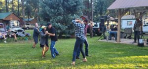 A group of people dance on a grassy area near a stage with a live band, creating a lively atmosphere reminiscent of an open-air gallery. Some are paired up, engaging in partner dances. Lawn chairs and small buildings are visible in the background, suggesting an outdoor community event.