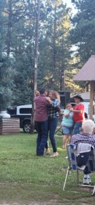 People are dancing outdoors at what appears to be a park or picnic area. Two couples are in the foreground: one in plaid shirts and jeans, while the second couple includes a man in a cowboy hat. Others are chatting or observing nearby, creating a lively scene reminiscent of an outdoor gallery.