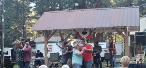 People are dancing in pairs outside under a pavilion with a small band playing music in the background. The pavilion, adorned with patriotic bunting and a metal roof, resembles an open-air gallery. Trees and vehicles create a festive, outdoor atmosphere that complements the lively scene.