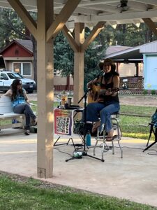 A musician wearing a hat plays an acoustic guitar and sings into a microphone under a pavilion. She is seated on a stool with a gallery-style QR code sign in front of her. Another person sits nearby, watching, with trees and cabins in the background.