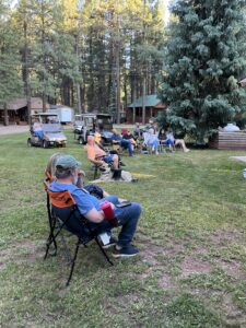 A group of people sitting on lawn chairs on a grassy area in a forested campground. Cabins and golf carts can be seen in the background like elements in an outdoor gallery. The group appears to be relaxing and socializing in the tranquil setting, with trees providing shade and creating a serene atmosphere.