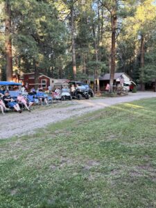 A group of people sit on benches and chairs in a forested campground, with a rustic cabin and an ATV visible in the background. The scene is set during the daytime, sunlight filtering through the trees like nature's gallery. An American flag hangs near the cabin.