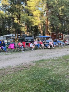 A group of people sitting on camping chairs arranged in rows, facing away from the camera. They are in a forested area with tall trees, with several parked vehicles and cabins in the background. The scene resembles an outdoor gallery as the group seems to be watching something out of frame.