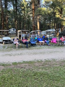 A group of people sits in folding chairs in a wooded area near RVs and golf carts, creating a natural gallery among the tall trees. They appear to be at a campsite, casually dressed, and engaged in conversation and relaxation.