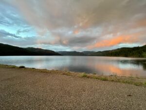 A serene lake surrounded by hills under a dramatic sky with a mix of blue, gray, and orange clouds. The calm water reflects the colors of the sky and distant tree-covered hills, creating a peaceful and picturesque scene reminiscent of an art gallery's finest display. Gravel beach in the foreground.
