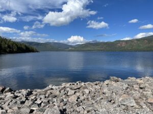 A serene lake surrounded by rocky shores is set against a backdrop of lush green hills and distant mountains under a sky filled with white, fluffy clouds. The view resembles a natural gallery, as the calm water reflects the vibrant blue of the sky.