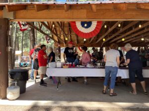 A group of people gathers under a wooden pavilion adorned with patriotic banners and string lights. They are standing around a long table with various dishes, engaging in conversation and helping themselves to the food, creating a scene reminiscent of fine art. Trees are visible in the background.