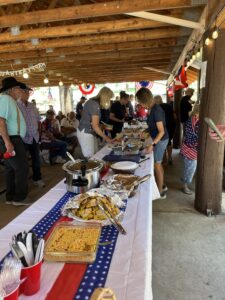 A group of people are gathered under a wooden pavilion with a red, white, and blue themed buffet table in the foreground. Various dishes, including a metal tray of food and plates of prepared items, are being served amid patriotic decorations hung around the pavilion, creating a lively gallery of celebration.
