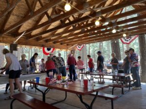 A group of people gather under a wooden pavilion decorated with red, white, and blue bunting. They are serving themselves food from a buffet table. Picnic tables with drinks and napkins are in the foreground like a festive gallery. String lights hang from the ceiling.