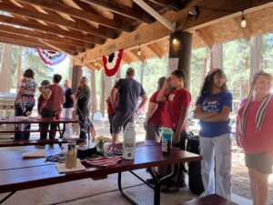 A group of people stands in line at an outdoor pavilion decorated with red, white, and blue bunting. Picnic tables display patriotic tablecloths and items like a lively gallery of Americana. The pavilion is open to a wooded area with tall trees in the background. Some people wear patriotic-themed clothing.