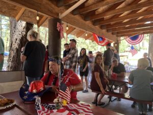 A group of people gathered under a wooden pavilion decorated with American flags and red, white, and blue banners. Some are seated at picnic tables, while others stand or wait in line. The atmosphere appears festive, like a lively gallery of patriotic decor and social interaction.
