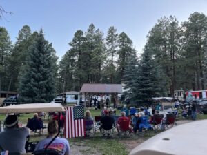A crowd of people sits in lawn chairs facing a small stage with musicians, set like an open-air gallery in a park surrounded by tall trees. An American flag is displayed prominently in the foreground, while some parked vehicles and a small building can be seen in the background.