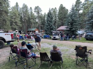 A group of people gathered at a campsite surrounded by tall trees. Some people are sitting in folding chairs, while others are standing or playing with a white dog. It looks like a nature gallery with several tents, cars, and a couple of rustic cabins visible in the background.