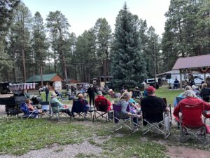A large group of people sit on folding chairs in a grassy, forested area, listening to music from a band on an outdoor stage. Cabins and trees form the backdrop, creating a natural gallery. Some individuals have blankets, and a dog is seen nearby. The atmosphere is relaxed.