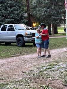 A couple, with a woman in a light blue shirt and shorts and a man in a red shirt and blue shorts, is standing close together, smiling outdoors. They are on a gravel path with a silver truck and trees in the background, creating a picturesque gallery-like scene. The man is wearing a black hat.