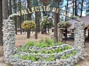 Entrance sign to Vallecito Resort, featuring a rustic stone archway with hanging flower baskets. Below the sign is a beautifully landscaped, stone-bordered garden filled with various green plants and flowers, resembling an outdoor gallery. Surrounding the area are tall trees and park-style buildings.