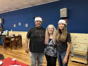 Three people are posing together indoors, smiling, with two of them wearing Santa hats and black t-shirts. The woman in the middle has long, light-colored hair and is wearing a floral top. The background resembles an art gallery with wooden paneling and decorative wall pieces.