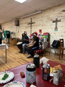 Two people are seated in a room decorated for Christmas, wearing Santa hats and drumming on djembes. In the foreground, a table holds food items and drinks. The wooden-paneled wall behind them features crosses and festive decorations, creating a cozy atmosphere reminiscent of an art gallery exhibition.