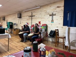 Two people wearing Santa hats play drums in a room with wooden walls. Behind them, holiday decorations and a cross are visible, lending an art gallery feel to the space. The room has tables with various items like cans, cups, and snacks. A blue curtain and some audio equipment are also present.