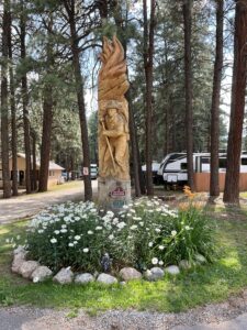 A wooden sculpture depicting a firefighter, surrounded by trees and flowers. The statue is carved from a tree trunk and stands among white and yellow flowers, enclosed by stones. To the left and right, there are recreational vehicles and a wooden structure visible, giving the scene an open-air gallery feel.