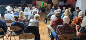 A crowd of people seated indoors in a community hall-style setting, attentively watching a stage performance that feels almost like an art exhibition. The audience consists mostly of older adults, and there are musicians performing at the front, with one person in a green outfit standing out.