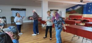 A group of elderly people is dancing and smiling in a community room with wooden floors, resembling a lively gallery. Tables with red tablecloths are arranged around the room, and a large mural of a landscape adorns the background wall.