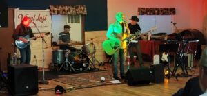 A four-man band performs on a small indoor stage, reminiscent of an art gallery exhibition. The members play electric guitar, drums, bass, and acoustic guitar. The lead singer, illuminated by a spotlight, stands at the forefront with a microphone. A sign reading "Vinyl Lust" is visible in the background.