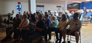 A group of people seated closely together, attending a gathering in a community hall that resembles a vibrant gallery. The room has wooden flooring, colorful murals on the walls, and various decorations. Some attendees are engaged in conversation while others appear to be listening attentively.