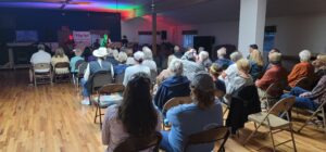 A small audience, mostly seniors, sit on chairs arranged in rows in a community hall with wooden flooring, facing a stage where a live band is playing under colorful lights. The room feels like an intimate gallery, cozy and well-lit, enhancing the atmosphere of this live exhibit.