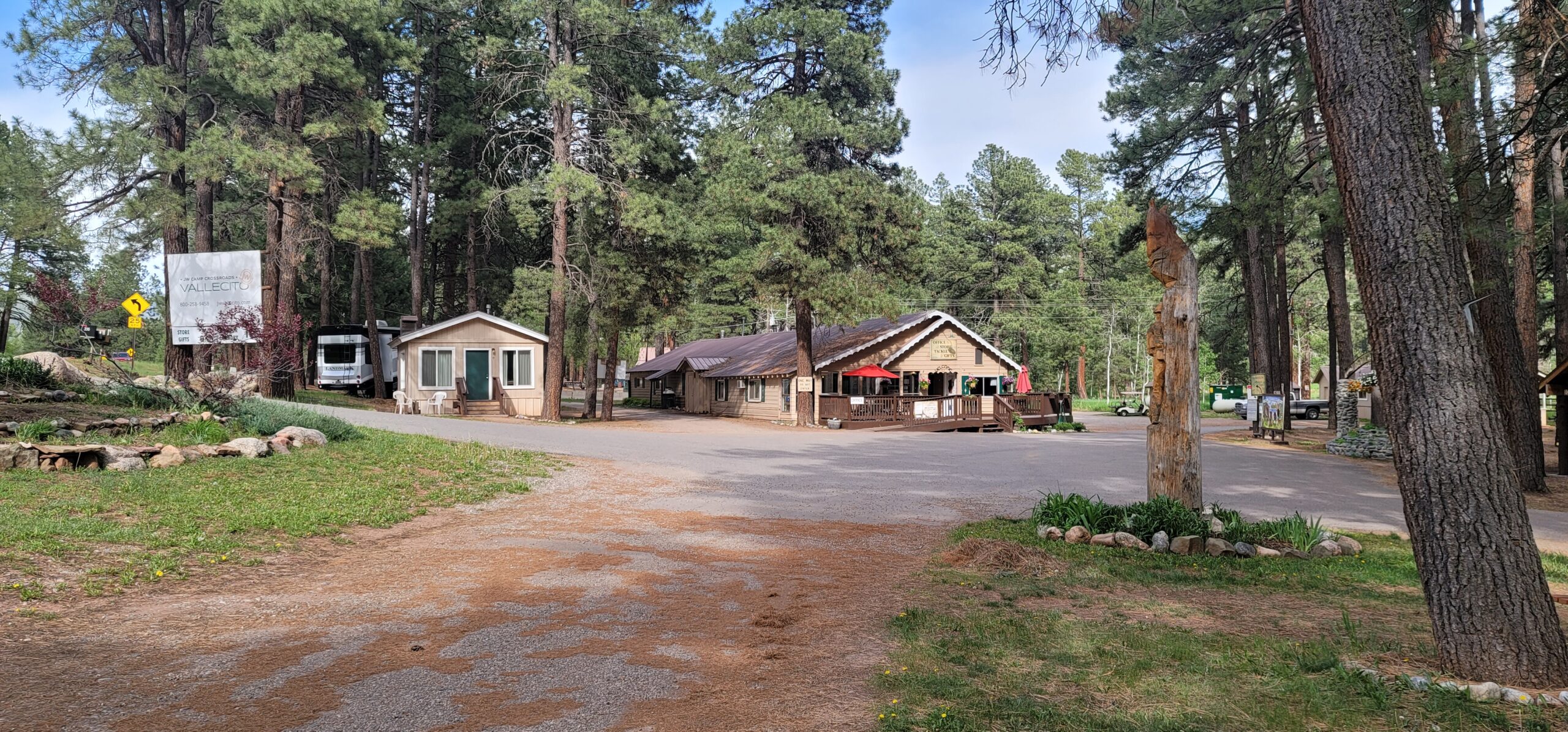 A rustic campground setting with two cabin-style buildings, surrounded by tall pine trees and offering both seasonal and monthly sites. A few cars and an RV are parked nearby. A wooden sculpture and a large sign nestle among the trees under a clear blue sky.