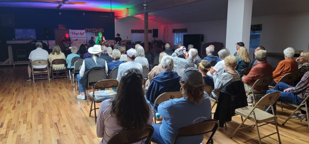 A group of people is seated in the Main Hall with wooden flooring, listening to a band performing on stage. The room is dimly lit with colorful stage lights, and most attendees sit in folding chairs, facing the musicians.